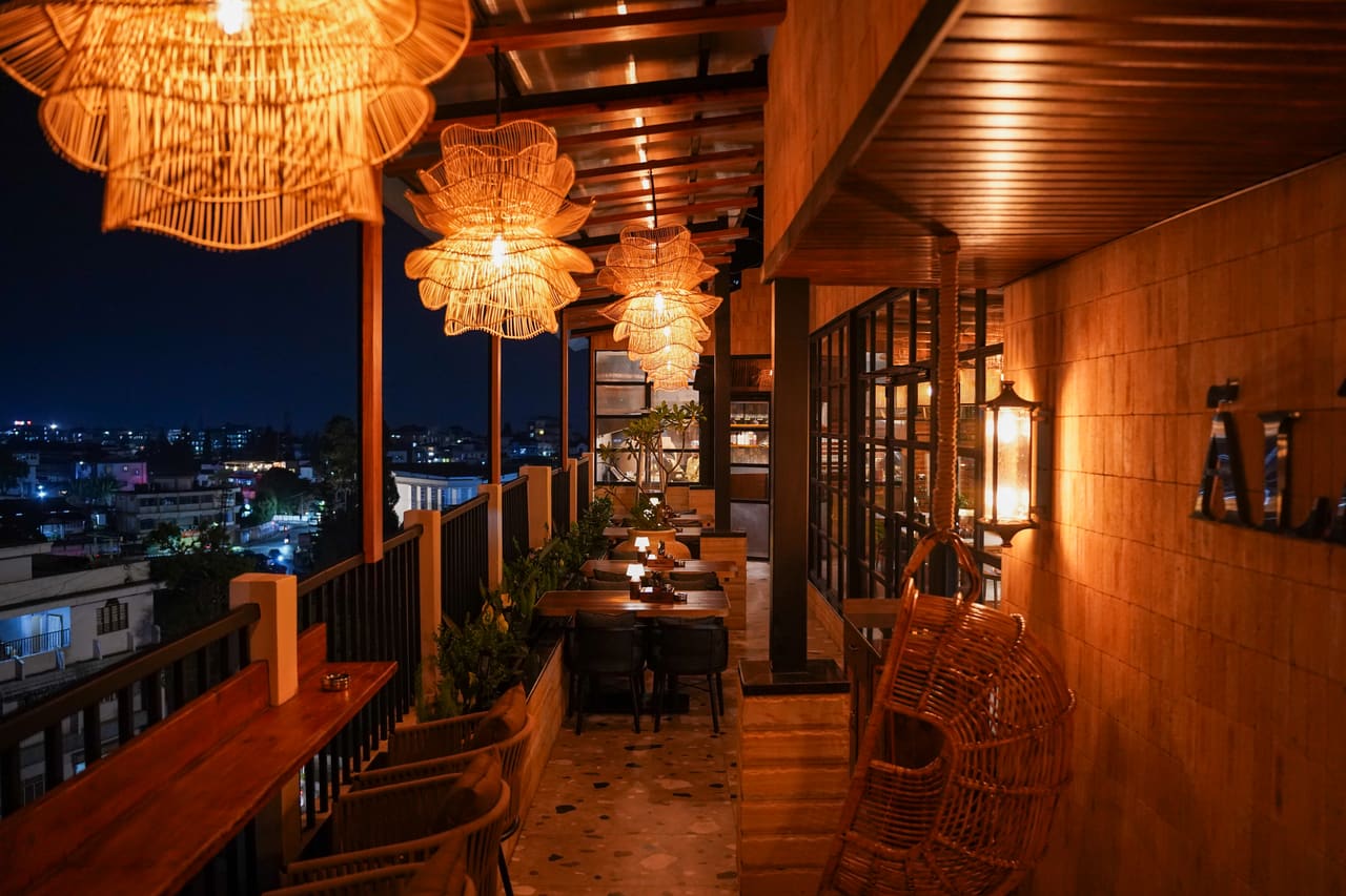 The Ālaya rooftop terrace at night, large rattan pendants glowing over the seating, with the lights of Shillong beyond the railing.